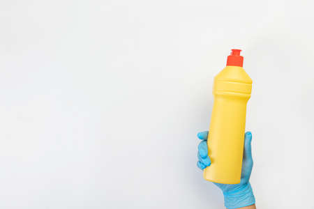 The hand of a cleaning lady in a blue rubber glove holds a bottle of cleaning agent on a gray background. Detergent for various surfaces in the kitchen, bathroom and other rooms.の写真素材
