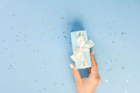 Flat lay of women's hands holding a gift wrapped and decorated with a bow on a blue background with silver sequins with copy spaceの写真素材