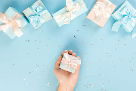 Flat lay of women's hands holding a gift wrapped and decorated with a bow on a blue background with silver sequins with copy space part of the table is surrounded by several festively wrapped giftsの写真素材
