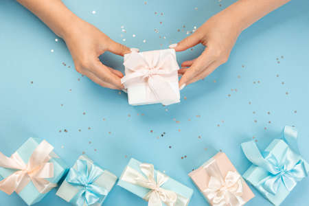 Flat lay of women's hands holding a gift wrapped and decorated with a bow on a blue background with...