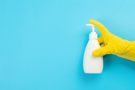 A hand in a yellow glove holds a white bottle of detergent on a blue background. Banner with copy...