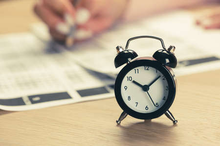 alarm clock with time running out. Students hands to take exams, write examination room with pencil holding on optical form of standardized test with answersの写真素材