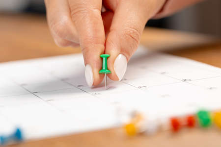 a woman's hand holds a green button to mark the date on a white calendar on the tableの写真素材