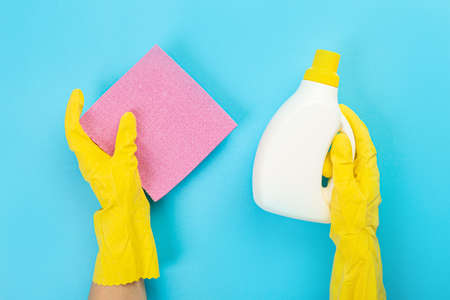 The hands of a housewife in yellow rubber protective gloves hold a bottle of household chemicals and a rag. Detergent for various surfaces in the kitchen, bathroom and other rooms. Light pastel blue background.の写真素材