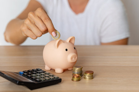 Close-up of a woman's hands put a euro coin in a piggy bank, next to a calculator on the table.correct investment. saving money in times of crisis and inflationの写真素材
