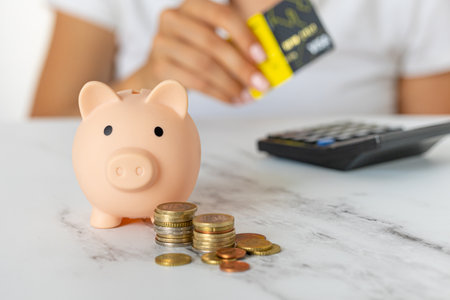 Close-up of a woman's hand with a credit card and a piggy bank, next to a calculator on the table. economy, crisis and inflation. Cost Managementの写真素材