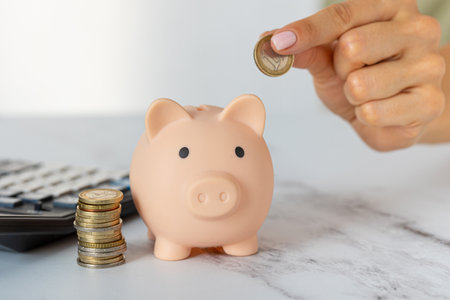 Close-up of a woman's hands put a euro coin in a piggy bank, next to a calculator on the table.correct investment. saving money in times of crisis and inflationの写真素材
