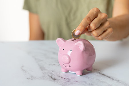 Close-up of a woman's hands putting a euro coin in a piggy bank, saving money as an investment in times of crisis and inflationの写真素材