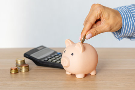 Close-up of a woman's hands put a euro coin in a piggy bank, next to a calculator on the table.correct investment. saving money in times of crisis and inflationの写真素材