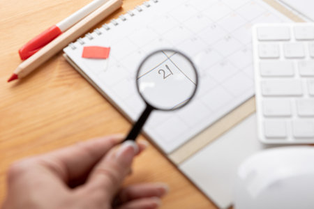 a hand with a magnifying glass examines the calendar and reminders for work from home online. glider on desktop with mouse and pen, keyboard. mark important plans in a diary at the office desk. 2023 calendar reminder concept.の写真素材