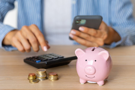 Close-up of a woman's hands put a euro coin in a piggy bank, next to a calculator on the table.correct investment. saving money in times of crisis and inflationの写真素材