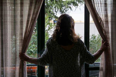 Rear view of a pretty young woman standing in the doorway on the balcony and looking outside into...