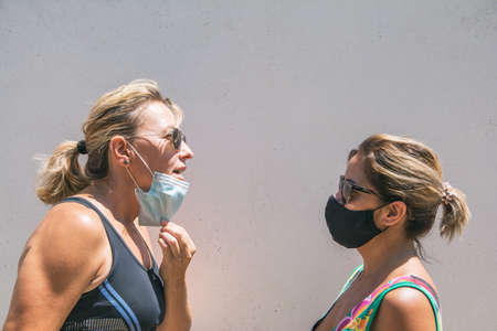 Young woman taking off protective mask to breathe and talk to friend. COVID-19. Two female friends...
