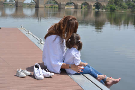 Mother and daughter enjoying a beautiful day sitting on a pier next to the river. Family conceptの写真素材