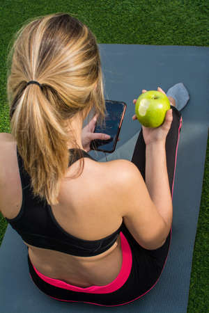 Young Woman Resting After Doing An Exercise Routine. She Is Using The Mobile Phone And Eating An...