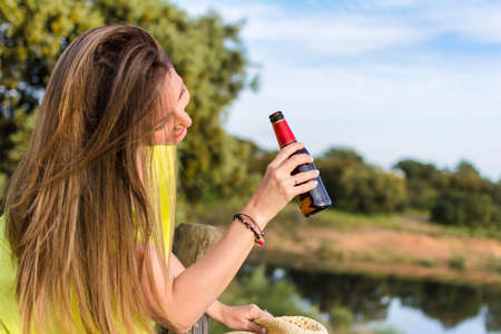 Beautiful Woman Drinking Beer At An Outdoor Campsite. Side View Of A Woman Drinking Beer From Bottles And Leaning On The Wooden Railing As She Relaxes At A Summer Campsite. Travel Concept.の写真素材