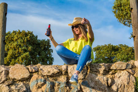 Beautiful Woman Sitting On A Camping Wall, Drinking A Bottle Of Beer While Watching The Sunset. Nature And Lifestyleの写真素材