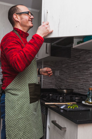 A man in a red fleece and a green apron cooking grilled artichokes and asparagus in a kitchen. He is opening a kitchen cupboard.の写真素材