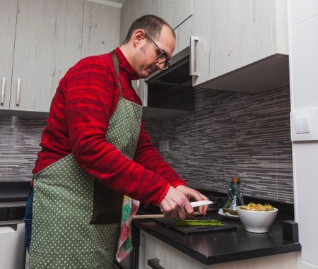 A man in a red fleece and a green apron cooking grilled artichokes and asparagus in a kitchen. He is cutting asparagus.の写真素材