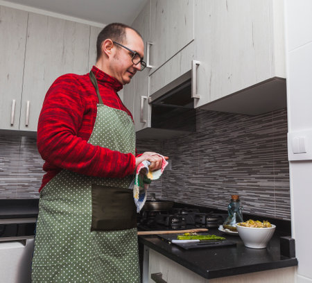 A man in a red fleece and a green apron cooking grilled artichokes and asparagus in a kitchen. He is drying his hands.の写真素材