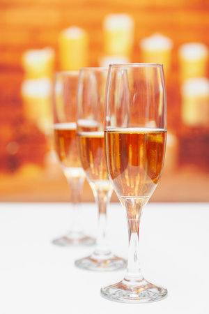 Three glasses of champagne on a white table, the first is in focus. The background is out of focus, one can sense that it is a fireplace with burning candles.の写真素材