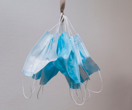 Many hanging surgical masks tied by the rubber bands with a cloth ribbon. In the background is a white wall.の写真素材