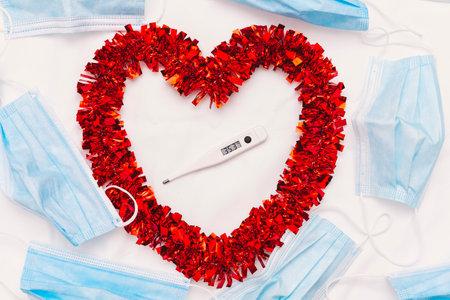Red decorative heart surrounded by blue surgical masks on a white cloth. In the center of the heart is a body thermometer that reads a temperature of 35.3 degrees Celsius.の写真素材