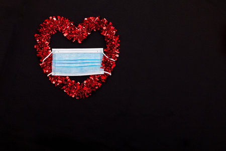 A red decorative heart with a blue surgical mask placed in the upper right corner of the image on a black fabricの写真素材