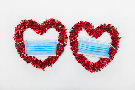 Two red decorative hearts with blue surgical mask placed on a white clothの写真素材
