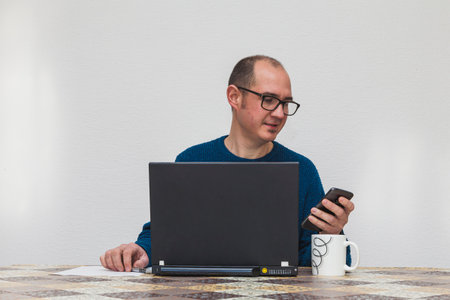 A bespectacled man dressed in a blue sweater using the touch screen of his smartphone in front of a laptop. On the table is a white cup, a sheet of paper, and a pen.の写真素材