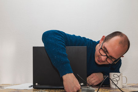 A man with glasses and a blue sweater is connecting the power cable of a laptop. On the table there is a white cup, a smartphone, a sheet of paper and a pen.の写真素材