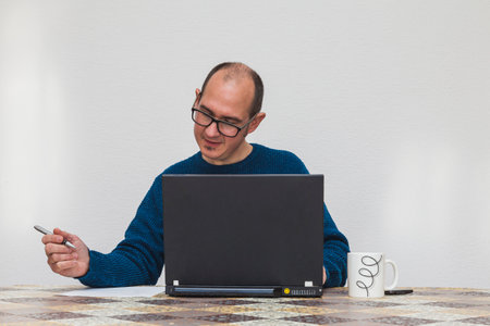 A bespectacled man dressed in a blue sweater drinking in front of a laptop. On the table there is a white mug, a mobile phone and a sheet of paper. In the background is a white wall.の写真素材
