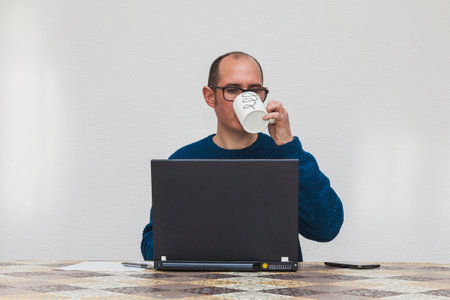 A bespectacled man dressed in a blue sweater drinking coffee from a white mug in front of a laptop. On the table there is a mobile phone, a sheet of paper, and a pen. In the background is a white wall.の写真素材