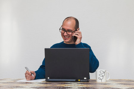 A man with glasses and in a blue sweater is talking with a smartphone in front of a laptop. On the table there is a white cup, a smartphone and a sheet of paper. He is holding a pen.の写真素材
