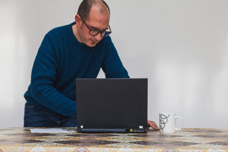 A man with glasses and a blue sweater is half sitting in front of a laptop. On the table there is a white cup, a smartphone, a sheet of paper and a pen.の写真素材