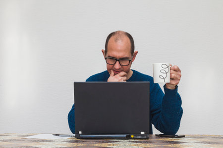 A man with glasses and in a blue sweater is pensive holding a white mug in front of a laptop. On the table there is a smartphone, a sheet of paper and a pen.の写真素材