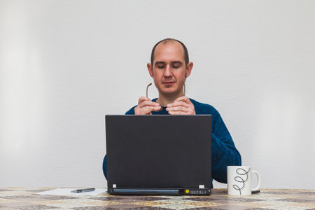 A man in a blue sweater is holding and looking at his glasses in front of a laptop. On the table there is a white cup, a smartphone, a sheet of paper and a pen.の写真素材