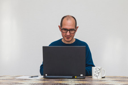 A man with glasses and in a blue sweater is typing on a laptop. On the table there is a white mug, a smartphone, a sheet of paper, and a pen.の写真素材