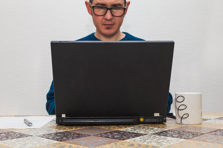 A man with glasses and in a blue sweater is typing on a laptop. On the table there is a white mug, a smartphone, a sheet of paper, and a pen.の写真素材