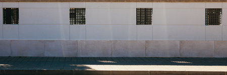 White exterior wall on a gray cobblestone sidewalk. In the wall there are four square windows with waffle-style metal grille. In the lower part of the wall there is a cladding of tiles.の写真素材