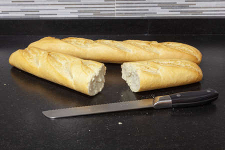 Two loaves of bread on a black granite bench in a kitchen. One of the bars is cut and next to it is the knife with which it was cut. You can see a little bit of the wall tiles.の写真素材