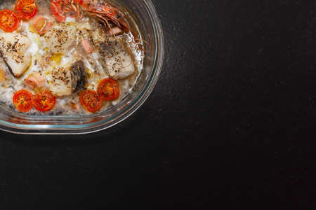 Glass tray with a recipe for baked cod with vegetables. There are fish, shrimp, cherry tomatoes, leek, onion, garlic, aromatic herbs and white wine. Part of the tray is hidden. There is copy space.の写真素材
