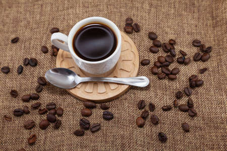 On a wooden plate there is a white cup with coffee and a metal spoon. They are placed on a sackcloth and there are coffee beans scattered around. It is seen from above.の写真素材