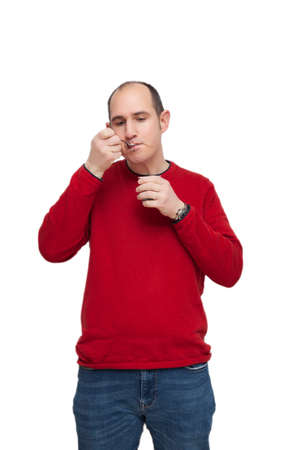 A bald young man wearing a red sweater is snacking on dairy with a metal spoon. He has the food in his mouth. The background is white.の写真素材