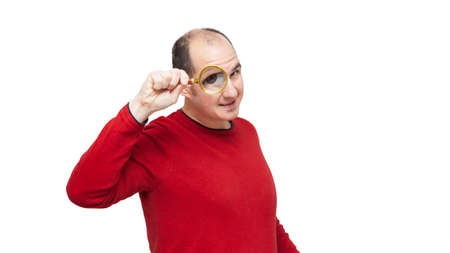 A bald young man wearing a red sweater is looking forward through a magnifying glass and his right eye is very large. The background is white.の写真素材