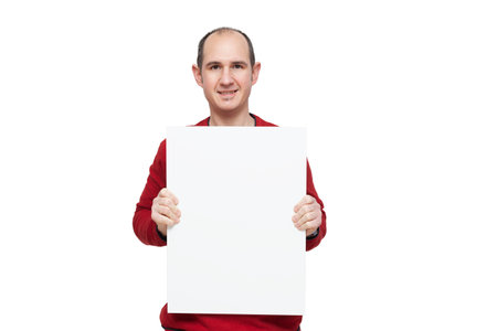A bald young man dressed in a red sweater is holding with his hands a blank poster in front of himself placed vertically. The background is white.の写真素材
