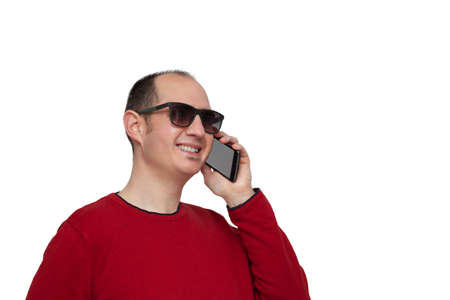 A bald young man dressed in a red sweater and sunglasses is holding his smartphone with his left hand next to his ear and is chatting with someone. The background is white.の写真素材