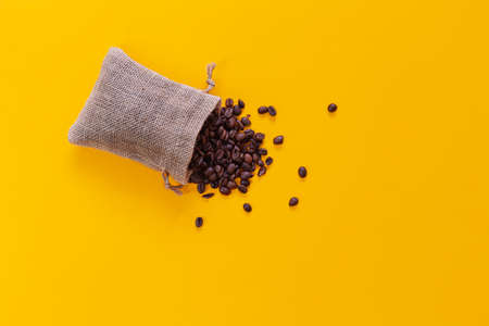 A small open sack is lying on a yellow background and coffee beans are coming out of it viewed from above. There is space for text or logos.の写真素材