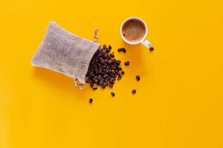 There is a small sack open and lying down from which coffee beans fall and scatter on a yellow background viewed from above. At the top is a white mug filled with coffee.の写真素材