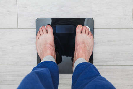 The feet of an unrecognizable man on top of an electronic scale where the weight is not visible. The man is wearing blue pajama pants and the floor is gray.の写真素材